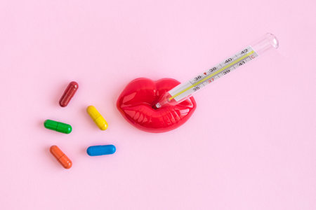Flat lay of plastic female lips with thermometer and colorful pills on pastel pink background...