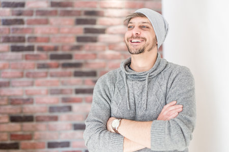 Young handsome smiling man with the beard in grey cap standing by the brick wall.