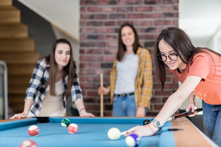 Cheerful female friends playing pool table billiard game at home.の写真素材