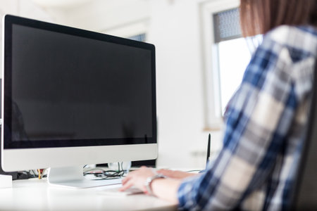 Rear view of female graphic designer sitting at the desk and using blank screen desktop computer at...