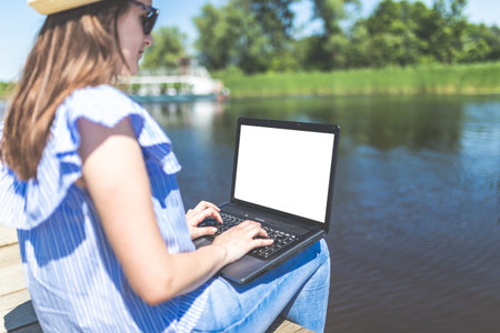 Side view of young woman sitting on pier and using blank screen laptop. Nature and technology concepts.の写真素材