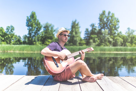 Modern fashionable man sitting on wooden pier and playing acoustic guitar with beautiful lake in the background. Music and vacation concepts.の写真素材