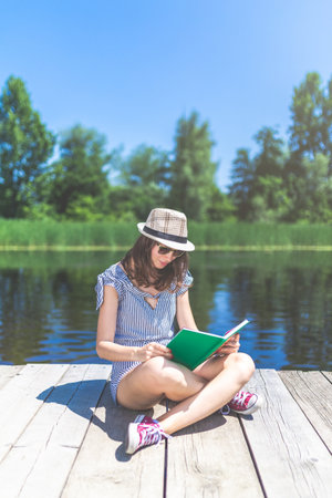 Fashionable woman relaxing on wooden dock and reading book.の写真素材