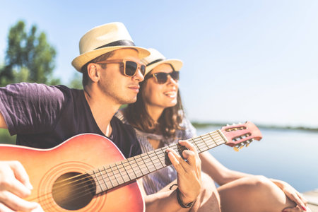 Beautiful fashionable couple with acoustic guitar against lake or river in the background. Summer and relationship concept.の写真素材