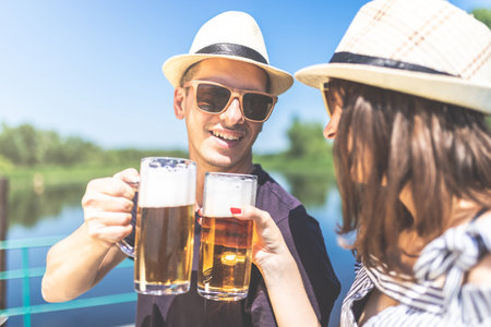 Young fashionable couple holding beer glasses against beautiful nature. Summer party concept.