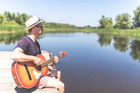 Side view of modern man sitting at pier edge, playing guitar and enjoying beautiful nature. Space for copy.の写真素材