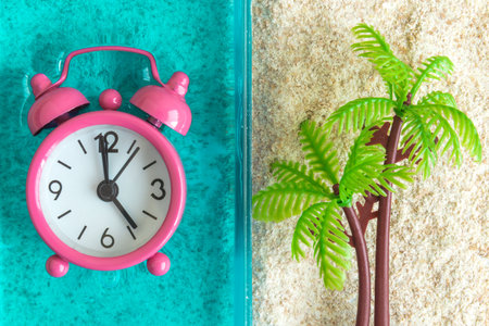 Flat lay of alarm clock in swimming pool or sea and palm tree in sand minimal creative summertime concept.の写真素材