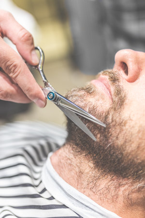 Close up of barber cutting beard of his client with scissors at barbershop.の写真素材