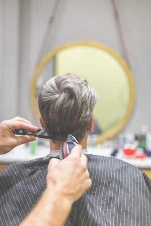 Rear view of professional barber styling hair of his client by using comb and clipper.