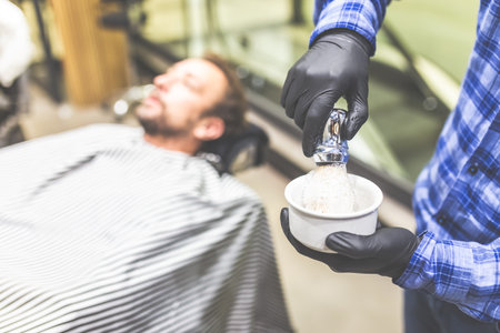 Close up of barber hands preparing foam by using shaving brush and bowl. Client in armchair in the...