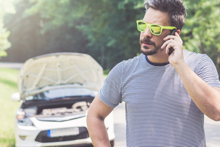 Young modern man making a call with road assistance service by using smartphone. Car with raised...