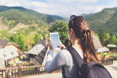 Female tourist holding blank screen digital tablet device against beautiful mountain village. Travel and technology concept.の写真素材