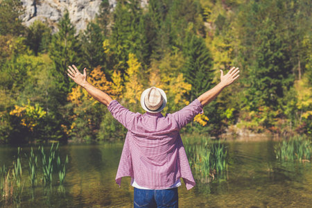 Rear view of modern young man with raised arms against beautiful lake and trees landscape. Travel and nature concepts.の写真素材