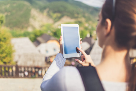 Rear view of beautiful young woman using tablet device near retro mountain village wooden bungalows.の写真素材