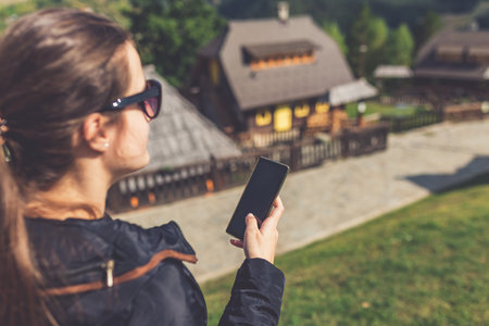 Beautiful young woman using smart phone near retro mountain village wooden bungalows.