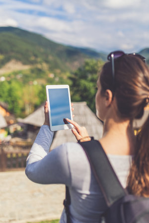 Female tourist holding blank screen digital tablet device against beautiful mountain village. Travel and technology concept.の写真素材
