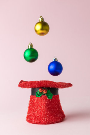 Colorful Christmas decoration baubles falling into hat against pastel pink background minimal...
