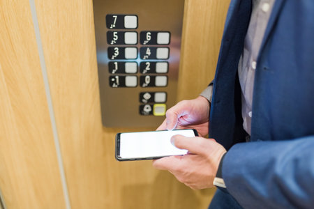 Businessman in elevator holding empty screen smartphone next to control panel.