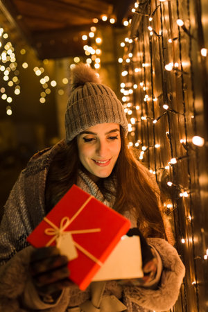Smiling woman opening christmas present gift box next to fence decorated with holiday lights on the street.の写真素材