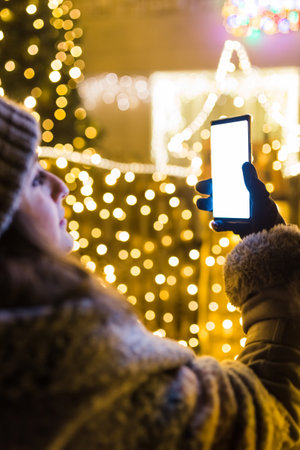 Woman using empty screen smart phone in front of house with new year christmas decoration lamps.の写真素材