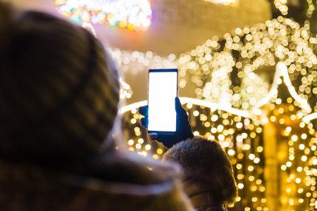 Rear view of young female holding blank screen mobile phone against house decorated with christmas lights.の写真素材