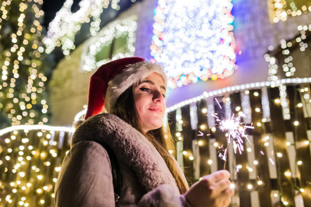 Portrait of beautiful woman wearing santa hat and holding sparkler in front of house decorated with christmas lights.の写真素材