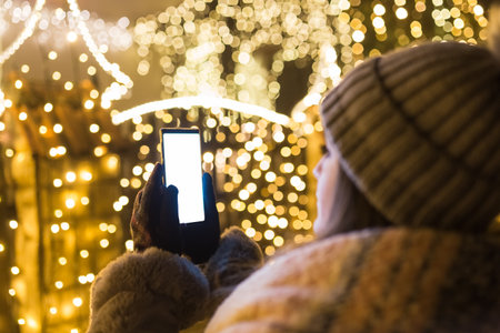 Woman using empty screen smart phone in front of house with new year christmas decoration lamps.の写真素材