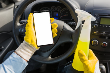 Close up of car wash worker holding blank screen smart phone and disinfectant spray bottle against car dashboard.の写真素材