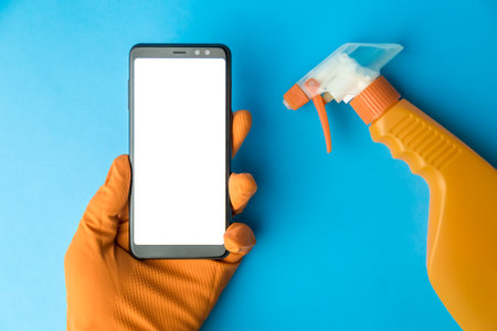 Hand wearing protective glove holding blank screen smartphone next to spray bottle against blue background minimal creative coronavirus or cleaning concept.の写真素材
