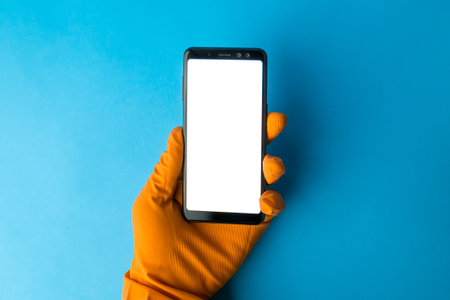Hand wearing protective glove holding blank screen smartphone against blue background minimal creative coronavirus or cleaning concept.の写真素材