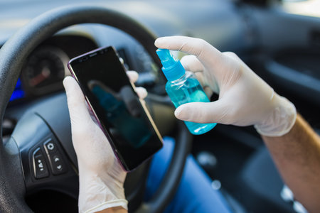 Close up of man driver wearing protective gloves and cleaning smartphone with alcohol spray in car. Germs or bacteria contamination.の写真素材