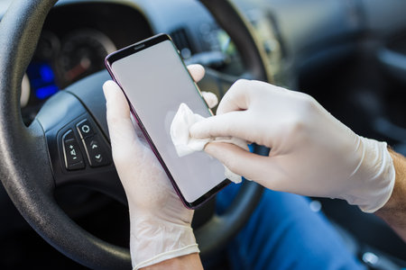 Close up of driver hands in surgical gloves wiping mobile phone with antibacterial wet wipe inside of car.の写真素材