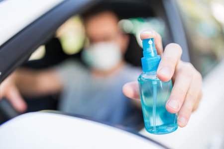 Portrait of man with surgical mask sitting in car and holding sprayer sanitizer. Hygiene concept.の写真素材