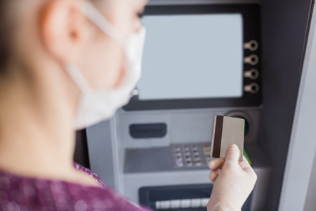 Rear view of young woman wearing protective surgical face mask and holding credit card in front of ATM machine to withdraw cash. Coronavirus concept.の写真素材