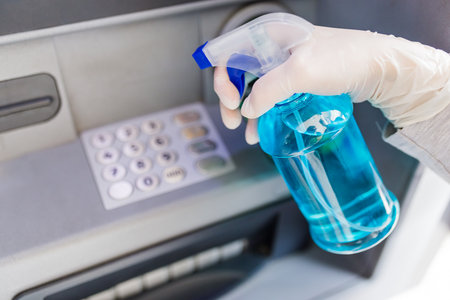 Close up of hand wearing surgical glove and holding bottle of cleaning alcohol to disinfect ATM numeric keyboard before withdrawing cash. Coronavirus concept.の写真素材