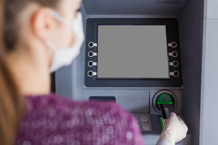 Rear view of young woman wearing protective surgical face mask and using ATM machine to withdraw cash. Coronavirus concept.の写真素材
