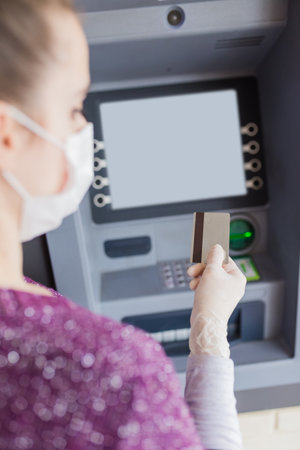 Rear view of young woman wearing protective surgical face mask and holding credit card in front of ATM machine to withdraw cash. Coronavirus concept.の写真素材