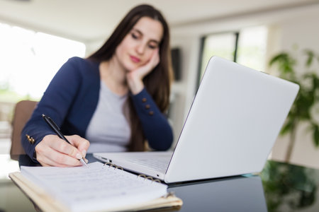 Portrait of young woman sitting at the desk in front of her laptop and taking notes. E-learning concept.の写真素材