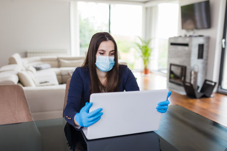 Young businesswoman working from home on laptop and wearing protective mask and gloves. Coronavirus quarantine concept.の写真素材