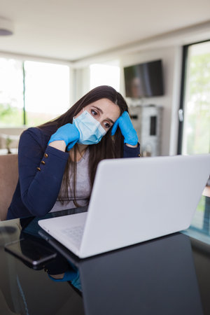 Young tired businesswoman with surgical mask and gloves working online from home, covid-19 isolation concept.の写真素材