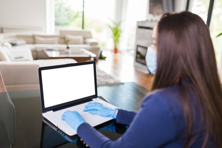 Rear view of woman wearing surgical gloves and mask while working on blank screen laptop. Work from home during coronavirus pandemic concept.の写真素材
