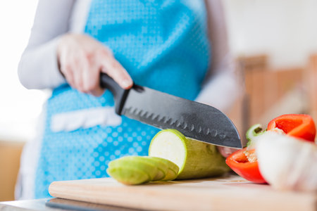 Close up of woman in apron slicing zucchini on cutting board in kitchen. Healthy meal preparation concept.の写真素材