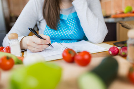Close up of woman in apron sitting at kitchen table and writing vegetable meal recipe.の写真素材