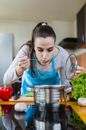 Portrait of young female tasting vegetable soup in kitchen. Healthy homemade meal concept.の写真素材