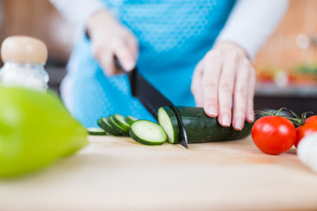 Close up of woman in apron slicing cucumber on cutting board in kitchen. Healthy meal preparation concept.の写真素材