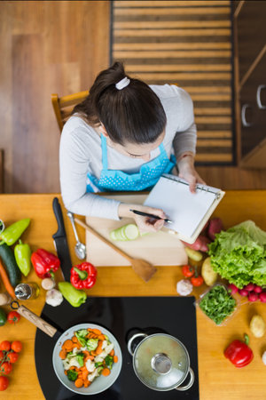 High angle view of woman in apron sitting at kitchen table and writing vegetable meal recipe.の写真素材