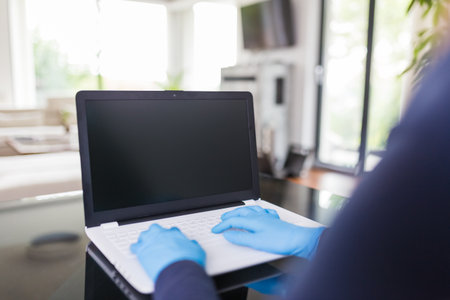 Rear view of woman wearing surgical gloves while working on blank screen laptop. Work from home during coronavirus pandemic concept.の写真素材