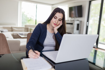 Portrait of young woman sitting at the desk in front of her laptop and taking notes. E-learning concept.の写真素材