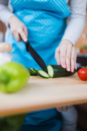 Close up of woman in apron slicing cucumber on cutting board in kitchen. Healthy meal preparation concept.の写真素材
