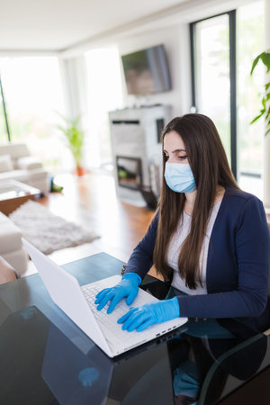 Woman wearing surgical gloves and mask while working on laptop. Work from home during coronavirus pandemic concept.の写真素材
